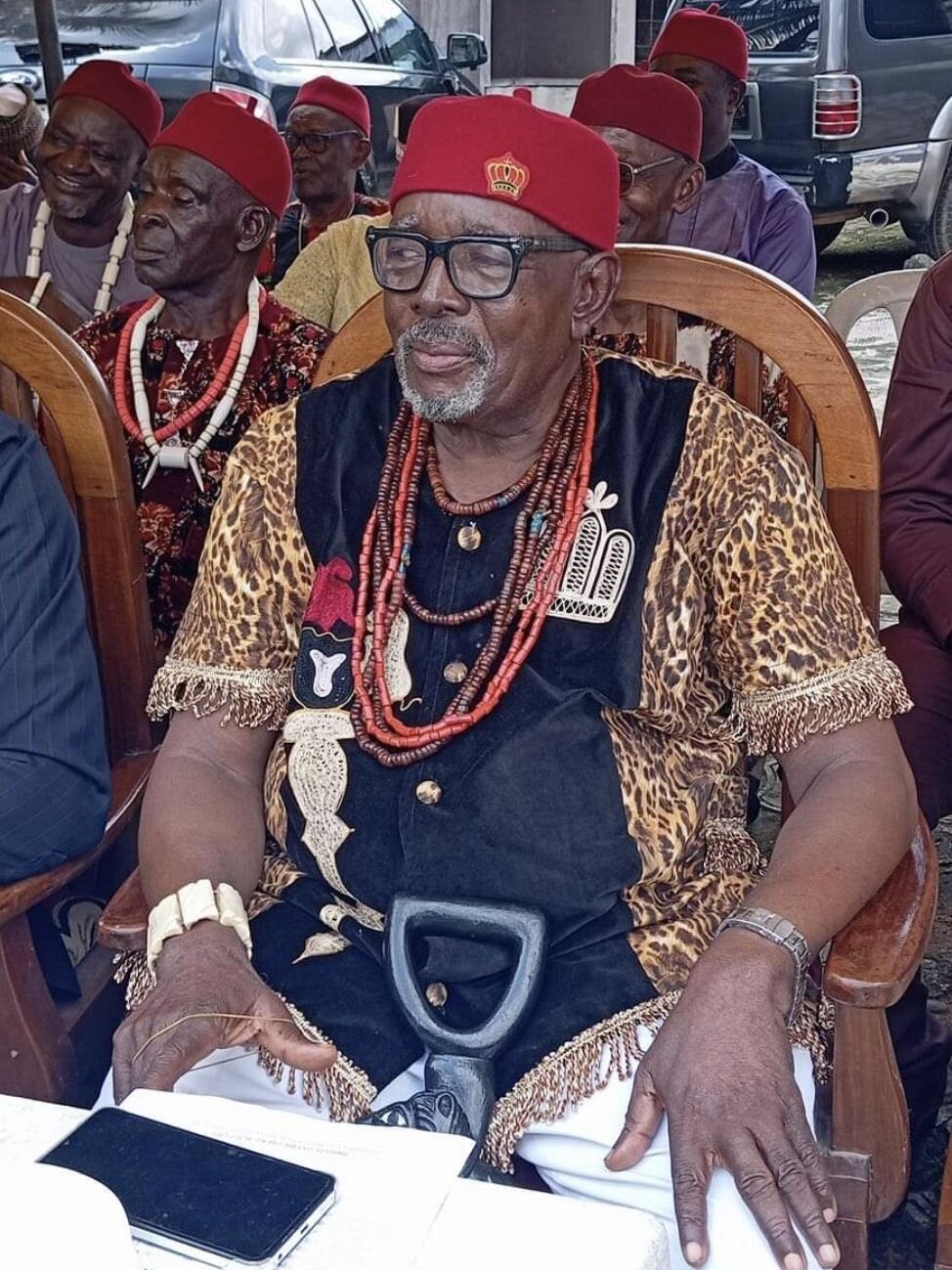 An elderly Ohaozara man dressed in traditional regalia and coral beads seated at a community function with other elders behind him.